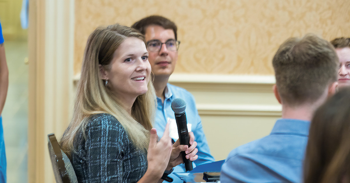A female sits at a table and holds a microphone while other people are also seated around the table.